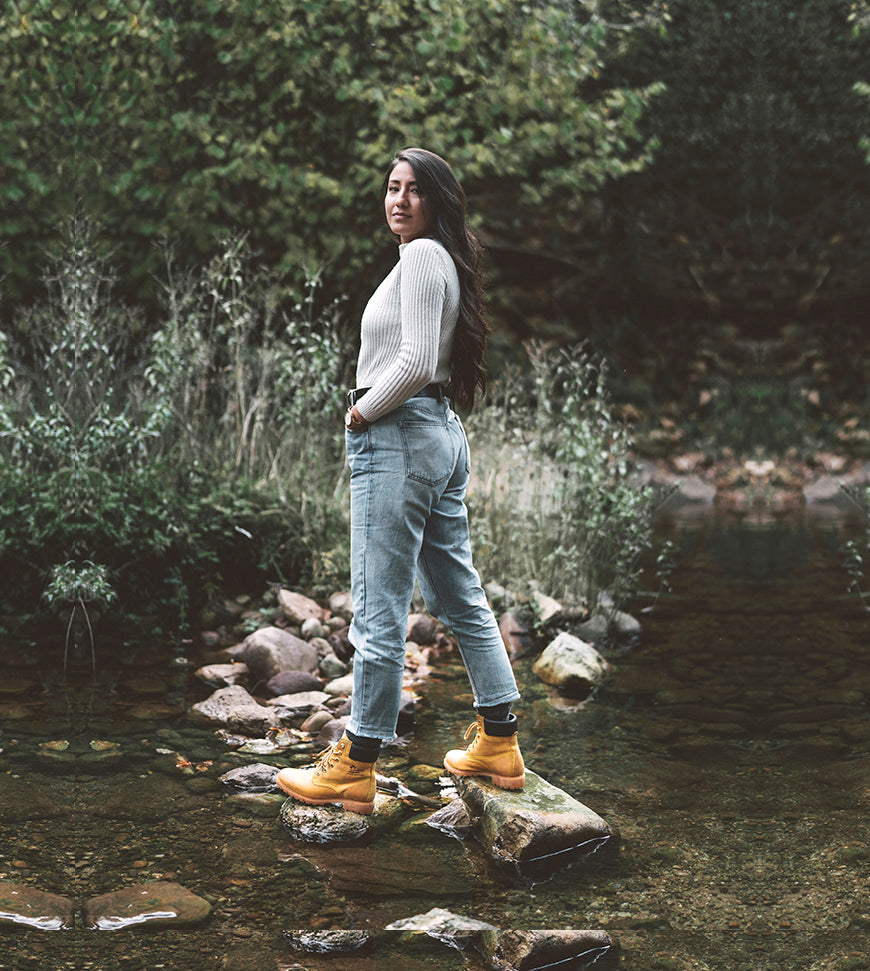 Chica con las botas icónicas de Panama Jack en unas piedras en mitad de un río de un bosque